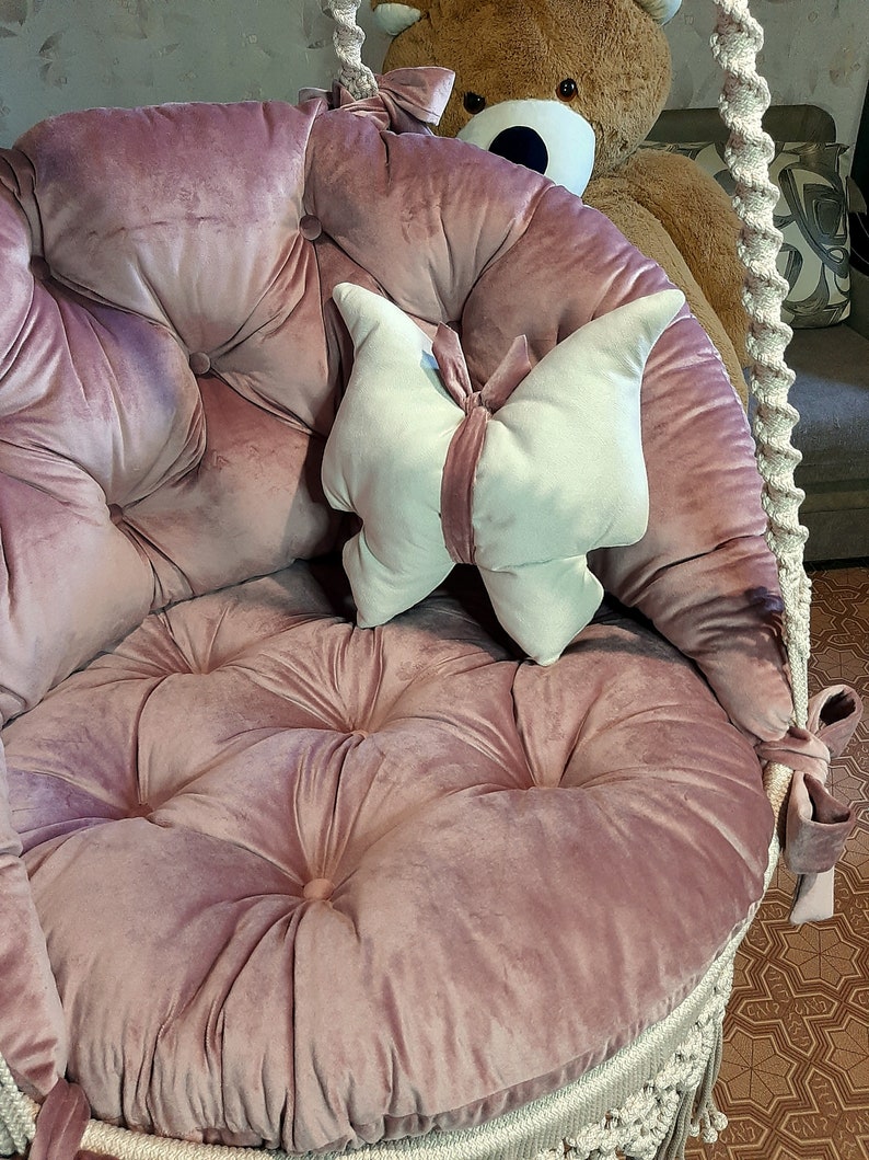 K&ouml;nnte beinhalten: Ein runder Schaukelstuhl in Staubrosa mit einer gesteppten Sitzfl&auml;che und R&uuml;ckenlehne. Ein schmetterlingsf&ouml;rmiges Kissen in Creme und Rosa liegt auf dem Sitz. Die Schaukel wird von dicken, gewebten Seilen gehalten.
