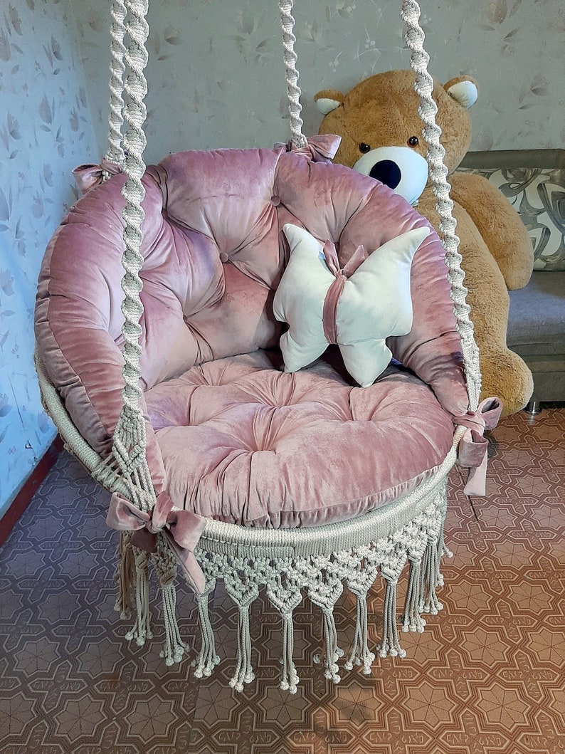 K&ouml;nnte beinhalten: Ein runder, rosa Samt-Schaukelstuhl mit Knopfheften und Makramee-Details. Der Stuhl hat einen wei&szlig;en Makramee-Fransenbesatz und ist an einem dicken, gedrehten Seil aufgeh&auml;ngt. Ein schmetterlingsf&ouml;rmiges Kissen liegt im Inneren.