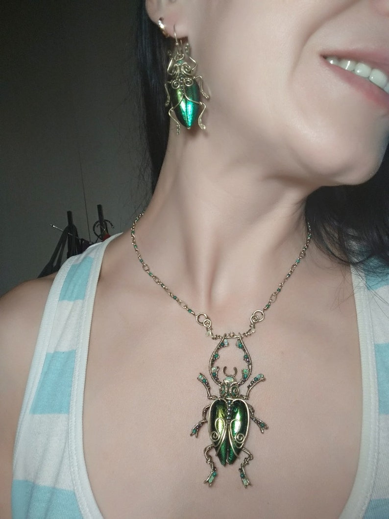 May include: A necklace and earrings set featuring beetle designs. The jewelry has a gold-colored wire frame with iridescent green beetle bodies. The necklace has a large pendant, and the earrings are smaller versions of the same design.
