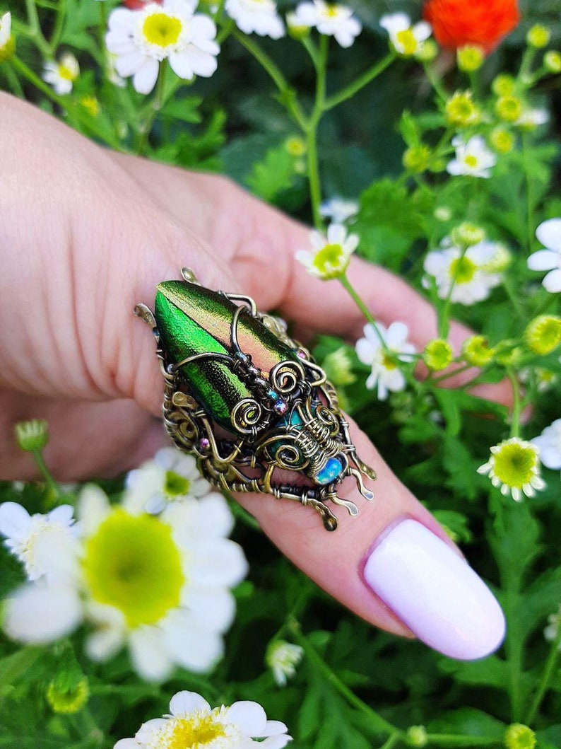 May include: A unique, handcrafted ring featuring a large, iridescent green beetle wing set in an ornate, antique brass-toned metal frame. The ring has intricate wirework and small, colorful accents, creating a statement piece. The ring is shown against a backdrop of green foliage and white flowers.