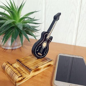 May include: A wooden phone stand featuring a black guitar design. The stand is crafted from wood with a burnt finish. A smartphone is resting on the stand.
