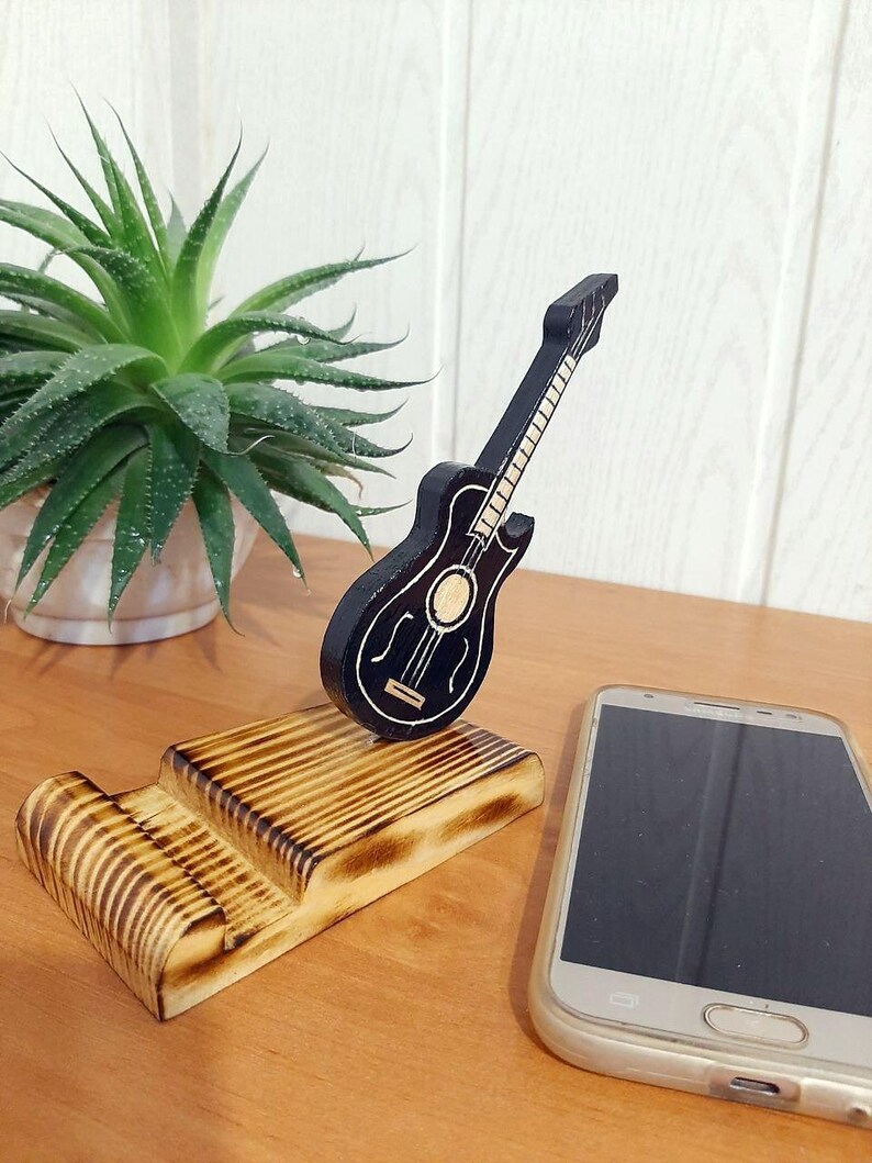 May include: A wooden phone stand featuring a black guitar design. The stand is crafted from wood with a burnt finish. A smartphone is resting on the stand.