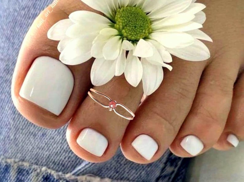 May include: Close-up of a foot with white nail polish and a toe ring with a pink gem. A white daisy flower rests on the foot. The image is set against a denim background.