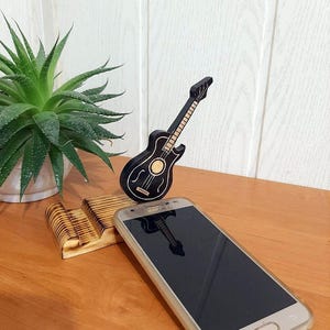 May include: A wooden phone stand featuring a carved guitar design. The stand is made of light brown wood and has a natural finish. A black smartphone is resting on the stand.