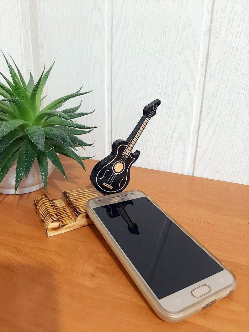 May include: A wooden phone stand featuring a carved guitar design. The stand is made of light brown wood and has a natural finish. A black smartphone is resting on the stand.