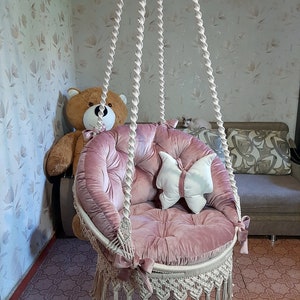 K&ouml;nnte beinhalten: Ein rosa Samt-H&auml;ngesessel mit Makramee-Fransen. Der Stuhl hat einen runden Sitz und R&uuml;cken mit getufteten Details. Ein wei&szlig;es, schmetterlingsf&ouml;rmiges Kissen liegt auf dem Sitz. Der Stuhl h&auml;ngt an einem dicken, gedrehten Seil.