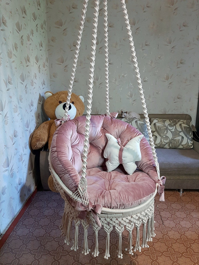 K&ouml;nnte beinhalten: Ein rosa Samt-H&auml;ngesessel mit Makramee-Fransen. Der Stuhl hat einen runden Sitz und R&uuml;cken mit getufteten Details. Ein wei&szlig;es, schmetterlingsf&ouml;rmiges Kissen liegt auf dem Sitz. Der Stuhl h&auml;ngt an einem dicken, gedrehten Seil.