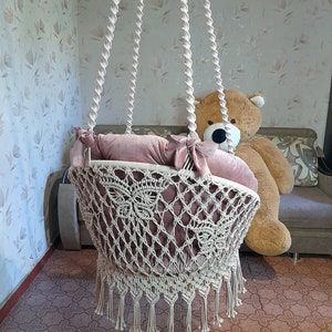 K&ouml;nnte beinhalten: Eine cremefarbene Makramee-Babyschaukel mit einem weichen, staubrosafarbenen Kissen. Die Schaukel wird mit dicken, gedrehten Seilen von der Decke abgeh&auml;ngt. Die Schaukel hat einen dekorativen Fransenbesatz am unteren Rand.