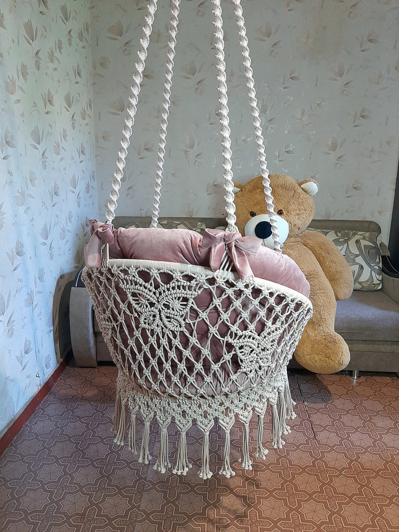 K&ouml;nnte beinhalten: Eine cremefarbene Makramee-Babyschaukel mit einem weichen, staubrosafarbenen Kissen. Die Schaukel wird mit dicken, gedrehten Seilen von der Decke abgeh&auml;ngt. Die Schaukel hat einen dekorativen Fransenbesatz am unteren Rand.