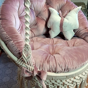 K&ouml;nnte beinhalten: Ein rosa Samt-H&auml;ngesessel mit rundem Sitz und R&uuml;ckenlehne. Der Stuhl hat ein Makramee-Design mit Fransen und einer dekorativen Schleife. Ein schmetterlingsf&ouml;rmiges Kissen liegt auf dem Sitz.