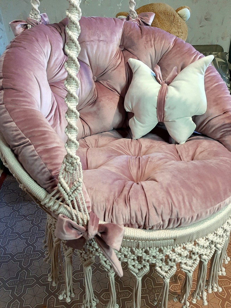 K&ouml;nnte beinhalten: Ein rosa Samt-H&auml;ngesessel mit rundem Sitz und R&uuml;ckenlehne. Der Stuhl hat ein Makramee-Design mit Fransen und einer dekorativen Schleife. Ein schmetterlingsf&ouml;rmiges Kissen liegt auf dem Sitz.