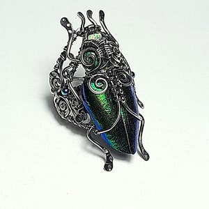 May include: A handcrafted, iridescent beetle pendant. The beetle's body is a vibrant mix of green, blue, and purple. It is encased in a detailed silver wire frame, with delicate antennae and legs. The pendant is set against a white background.