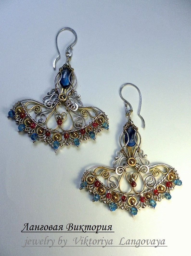 May include: A pair of silver and gold earrings with blue and red gemstones. The earrings are in the shape of a fan and have a delicate, intricate design. The earrings are labeled "jewelry by Viktoriya Langovaya".