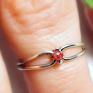 May include: A silver ring with a split band design, featuring a small, round, red gemstone at the center. The ring is shown on a finger, highlighting the delicate details of the jewelry.