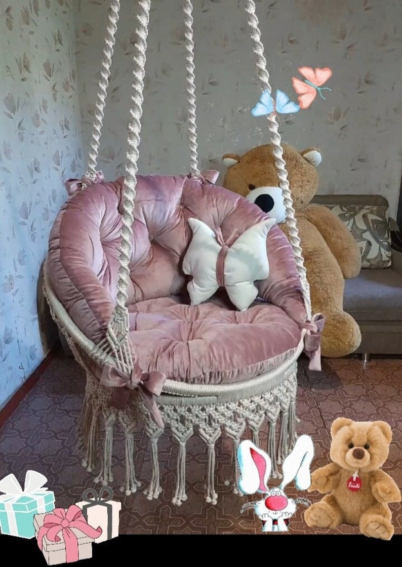 K&ouml;nnte beinhalten: Ein rosa Samt-H&auml;ngesessel mit Makramee-Details und einem kissenf&ouml;rmigen Schmetterling. Der Stuhl wird von dicken, gewebten Seilen gehalten. Ein gro&szlig;er Teddyb&auml;r sitzt hinter dem Stuhl.