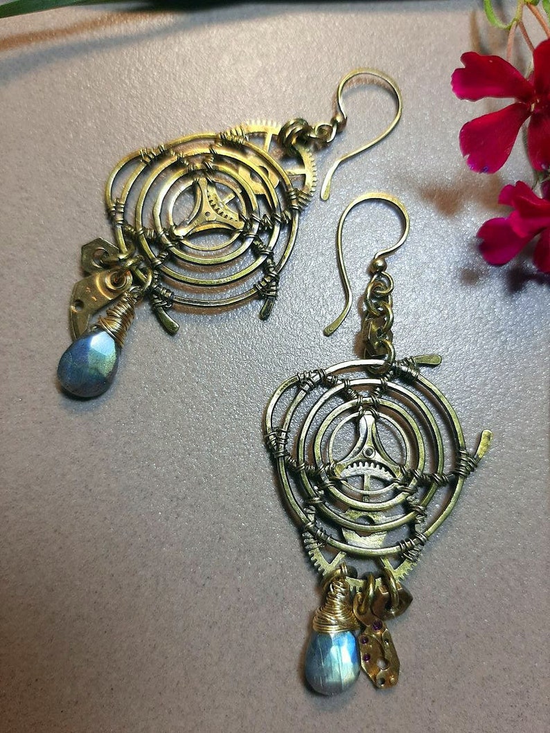 Może przedstawiać: Para złotych kolczyk&oacute;w w stylu steampunk. Każdy kolczyk ma okrągły wz&oacute;r z kołami zębatymi i wiszącym od dołu niebieskim kamieniem szlachetnym w kształcie łzy.