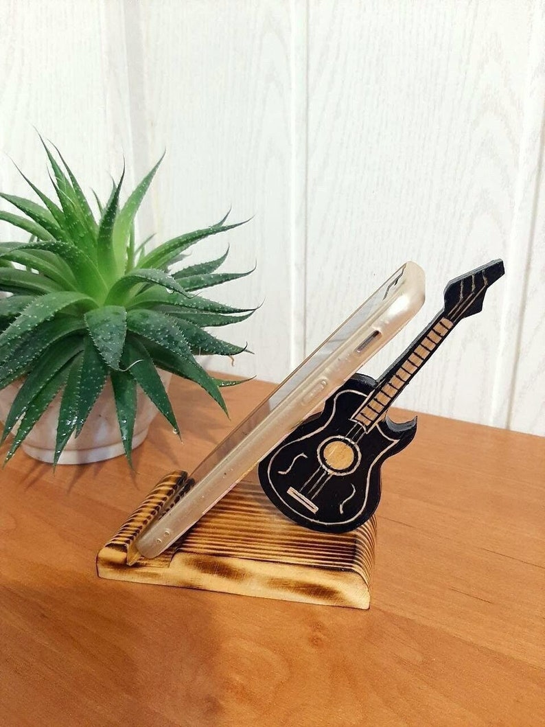 May include: A wooden phone stand featuring a carved guitar design. The stand is made of light brown wood with a dark brown stain. The guitar is black with white strings.