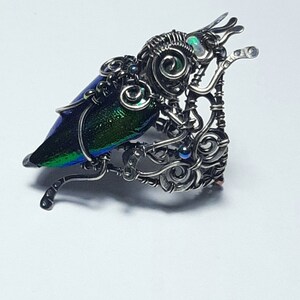 May include: A handcrafted, silver-toned metal sculpture of a beetle. The beetle's wing is iridescent with shades of blue and green. The sculpture has intricate wire detailing and small, blue accent stones.