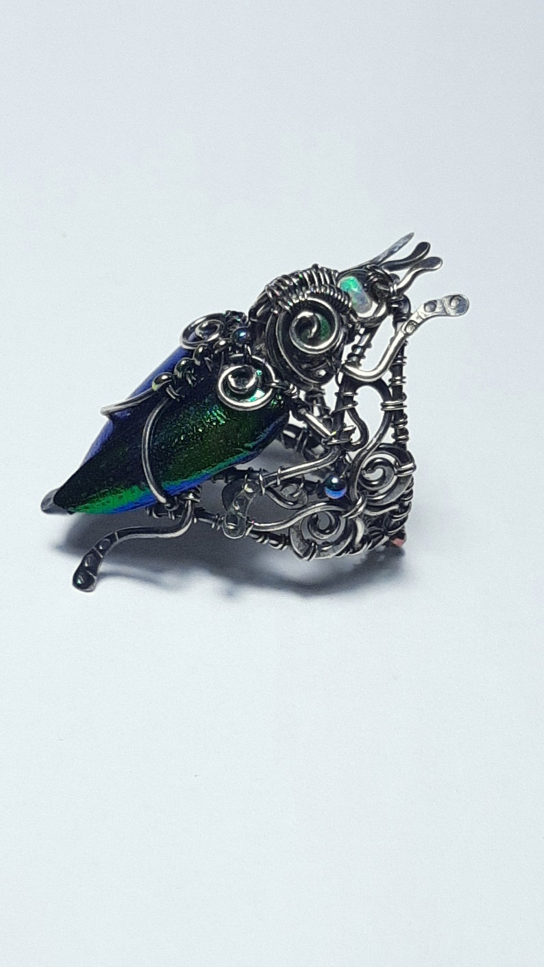 May include: A handcrafted, silver-toned metal sculpture of a beetle. The beetle's wing is iridescent with shades of blue and green. The sculpture has intricate wire detailing and small, blue accent stones.