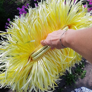 May include: A large, handmade yellow and white decorative item resembling a flower. The item is held by a person's hand, with a silver bracelet visible. The background includes green foliage and purple flowers, suggesting an outdoor setting.