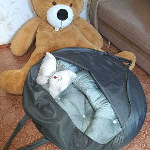 K&ouml;nnte beinhalten: Ein gro&szlig;er brauner Teddyb&auml;r mit wei&szlig;er Schnauze und schwarzen Augen sitzt in der N&auml;he eines dunkelgrauen Haustierbettes. Das Bett ist offen und zeigt einen hellblauen Innenraum mit wei&szlig;en Kn&ouml;pfen und einem wei&szlig;en Kissen.