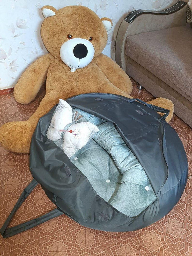 K&ouml;nnte beinhalten: Ein gro&szlig;er brauner Teddyb&auml;r mit wei&szlig;er Schnauze und schwarzen Augen sitzt in der N&auml;he eines dunkelgrauen Haustierbettes. Das Bett ist offen und zeigt einen hellblauen Innenraum mit wei&szlig;en Kn&ouml;pfen und einem wei&szlig;en Kissen.