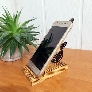 May include: A wooden phone stand with a guitar design. The stand is made of light brown wood and has a dark brown guitar design on the side. A gold smartphone is resting in the stand.