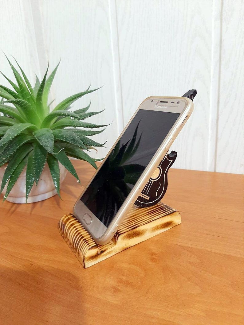 May include: A wooden phone stand with a guitar design. The stand is made of light brown wood and has a dark brown guitar design on the side. A gold smartphone is resting in the stand.