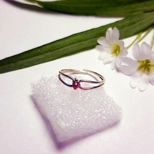 May include: A silver ring with a small, red gemstone at the center. The ring is set on a white, textured surface, with green leaves and small white flowers in the background. The ring has a double-band design.