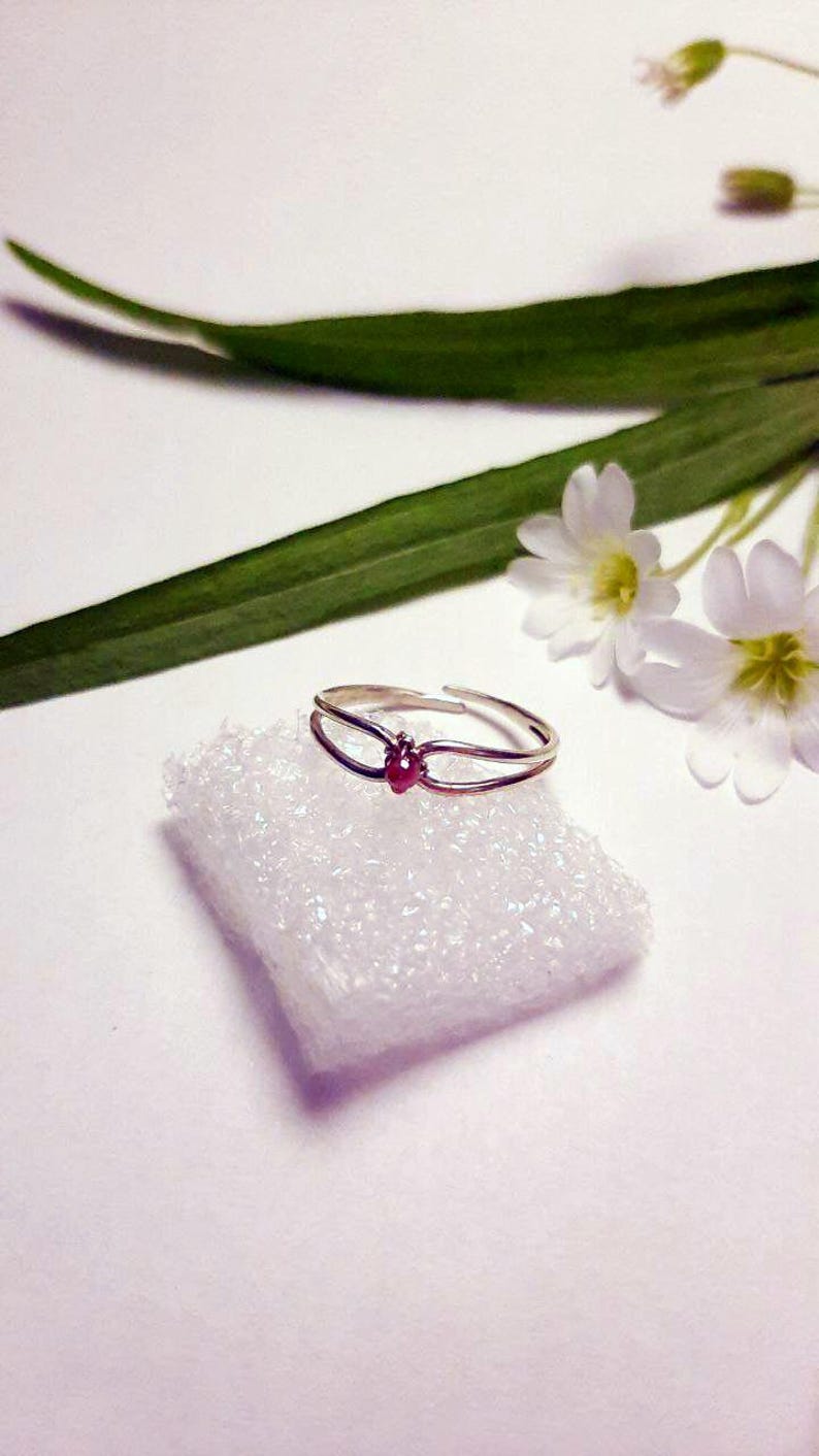 May include: A silver ring with a small, red gemstone at the center. The ring is set on a white, textured surface, with green leaves and small white flowers in the background. The ring has a double-band design.