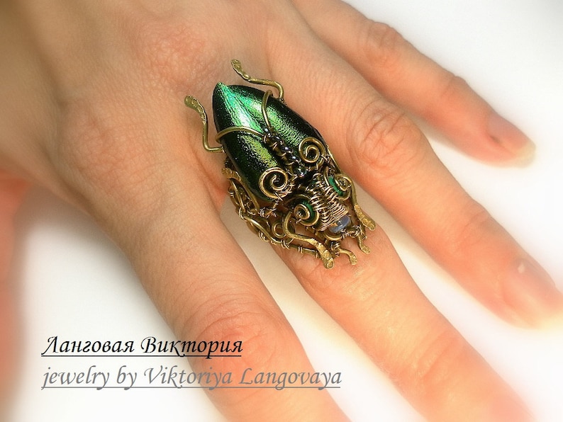 May include: A unique, handcrafted ring featuring a detailed beetle design. The beetle's body is a vibrant green, complemented by intricate wirework in gold tones. The ring's design includes small, decorative elements and is worn on a finger.