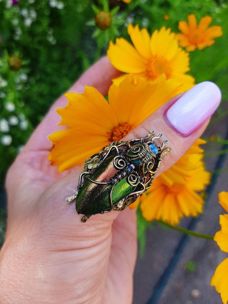 May include: A handcrafted ring featuring a beetle design. The ring showcases two iridescent green beetle wings, intricately wrapped in bronze-colored wire. Small blue and purple beads add accents. The ring is held near bright yellow flowers.