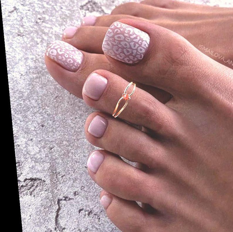 May include: Close-up of a foot with a gold toe ring and pink nail polish. Two toenails have a white leopard print design. The other nails are painted a solid light pink color.