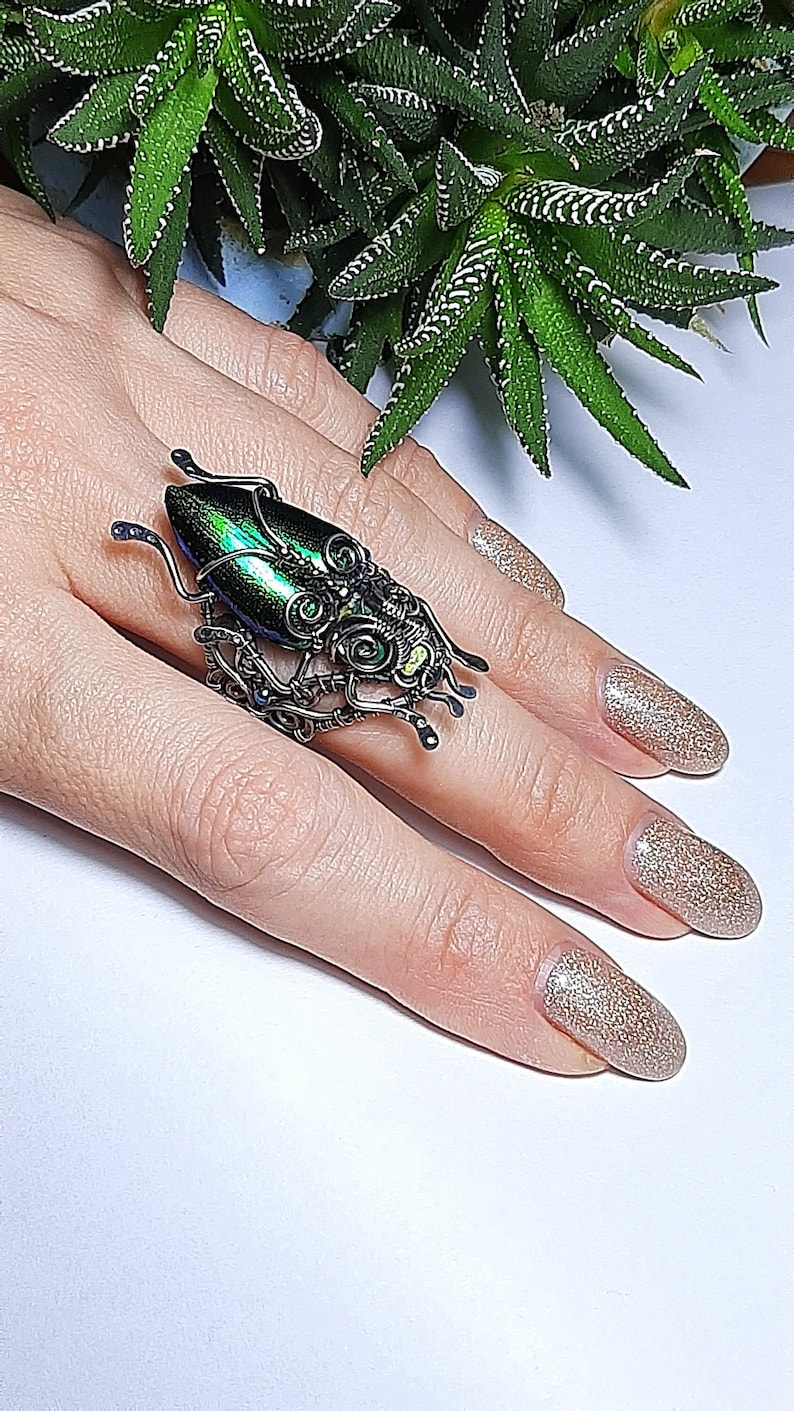 May include: A unique, handcrafted ring featuring a beetle design. The ring showcases a metallic green and black beetle body, accented with intricate wire detailing. The ring is a statement piece, perfect for adding a touch of nature-inspired artistry to any outfit.
