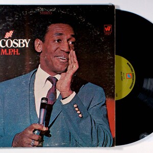 May include: Album cover for Bill Cosby's "200 M.P.H." record. The cover features a photo of Bill Cosby in a blue suit, holding a microphone. The album is a stereo record. The record is black.