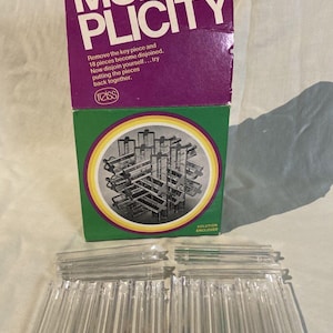 May include: A box with the text "MULTI-PLICITY" on the front. The box is purple and green and shows a 3D puzzle made of clear plastic pieces. The text "SOLUTION ENCLOSED" is on the bottom of the box. The box is sitting on a white surface with 18 clear plastic pieces.