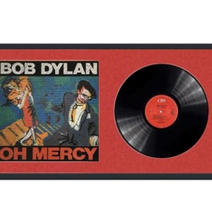 May include: A framed record album with a black vinyl record. The album cover features a colorful abstract design with the text "BOB DYLAN OH MERCY".