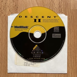 May include: A yellow and black Descent II CD-ROM for Macintosh computers, with the MacAttack logo. The disc is in a white paper sleeve, with the Aztech New Media logo and contact information visible. The disc is labeled "Compact Disc Data Storage".
