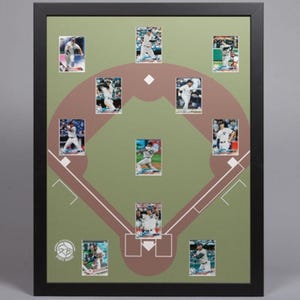 May include: A black framed display featuring ten baseball trading cards arranged on a green mat board printed with a brown baseball diamond graphic. The cards show various players in action poses. A circular logo is visible in the bottom left corner.