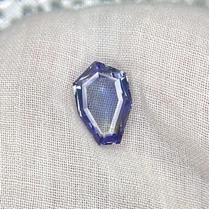 May include: A faceted, purple gemstone with a unique, geometric shape. The gemstone is set against a white fabric background.