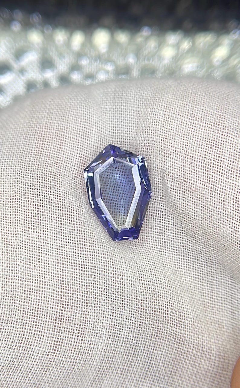 May include: A faceted, purple gemstone with a unique, geometric shape. The gemstone is set against a white fabric background.