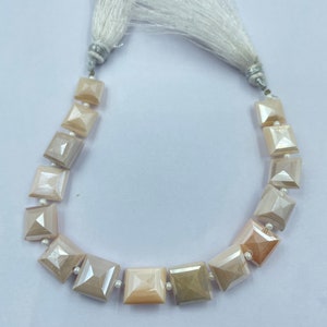 Puede incluir: Un collar con cuentas cuadradas e iridiscentes en tonos beige y melocotón, ensartadas con pequeñas perlas blancas. El collar está rematado con detalles de borlas blancas en cada extremo, creando un diseño delicado y elegante.