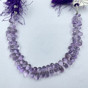 May include: A necklace featuring strands of faceted, teardrop-shaped amethyst beads. The beads graduate in size and are strung on a thin, clear cord. The necklace is accented with purple tassel fringe at each end.