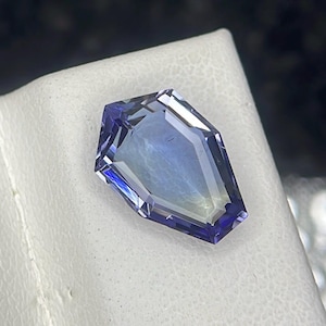 May include: A faceted, purple gemstone with a unique, coffin-shaped cut. The gemstone is set against a white background.