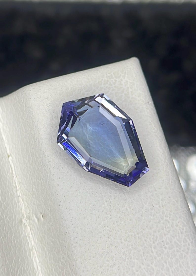 May include: A faceted, purple gemstone with a unique, coffin-shaped cut. The gemstone is set against a white background.