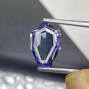 May include: A purple gemstone with a faceted, geometric shape. The gemstone is held by tweezers.