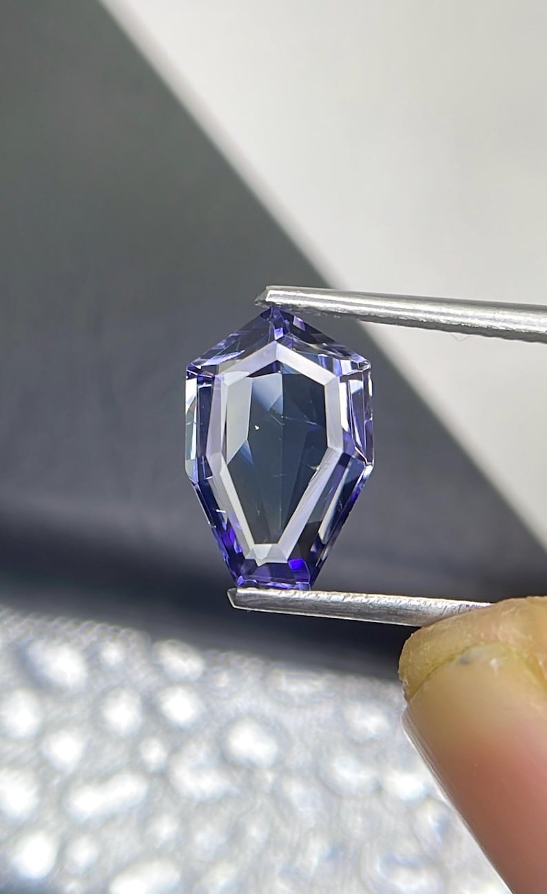 May include: A purple gemstone with a faceted, geometric shape. The gemstone is held by tweezers.