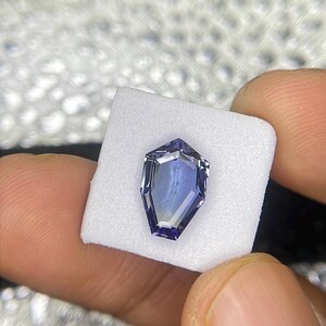 May include: A purple gemstone with a faceted, octagonal shape. The gemstone is set on a white foam pad.