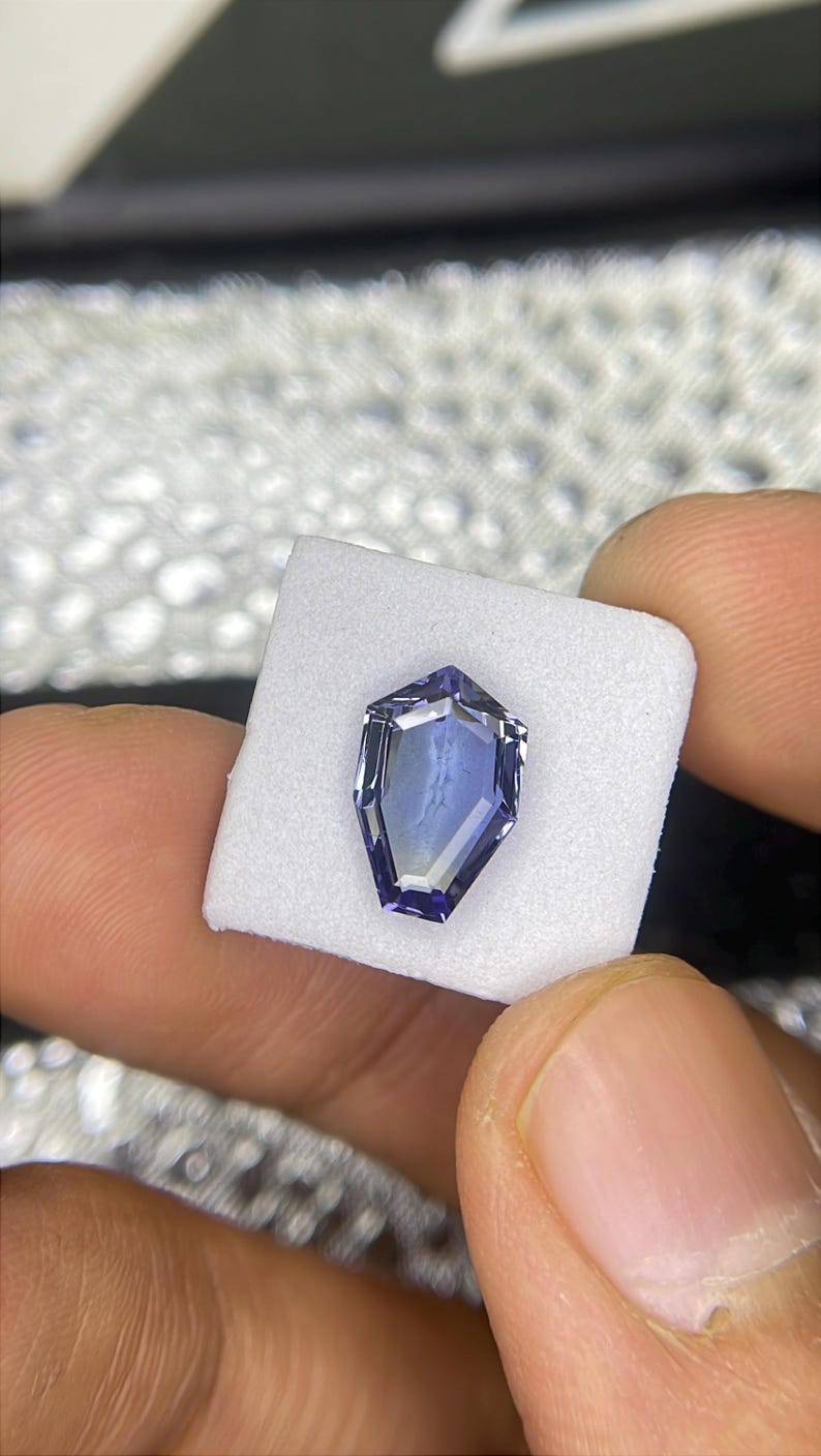 May include: A purple gemstone with a faceted, octagonal shape. The gemstone is set on a white foam pad.