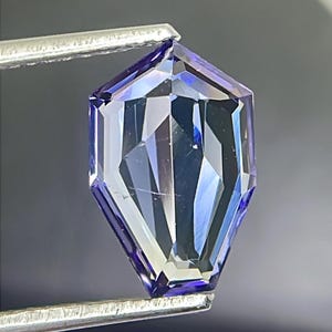 May include: A faceted, purple gemstone with a hexagonal shape, held by tweezers against a grey background.